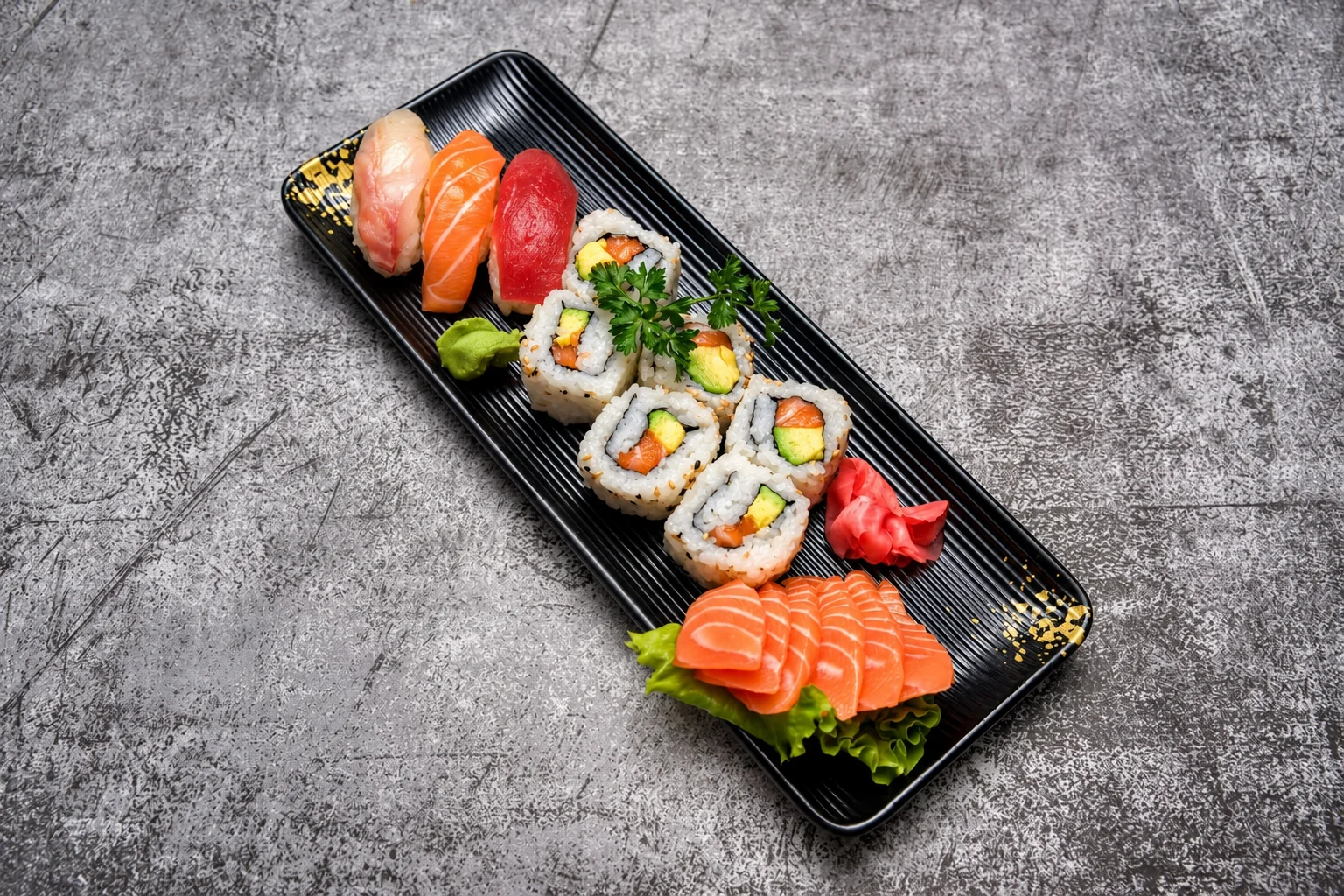 Nigiri, california rolls and salmon sashimi on a black rectangular plate, grey concrete background.