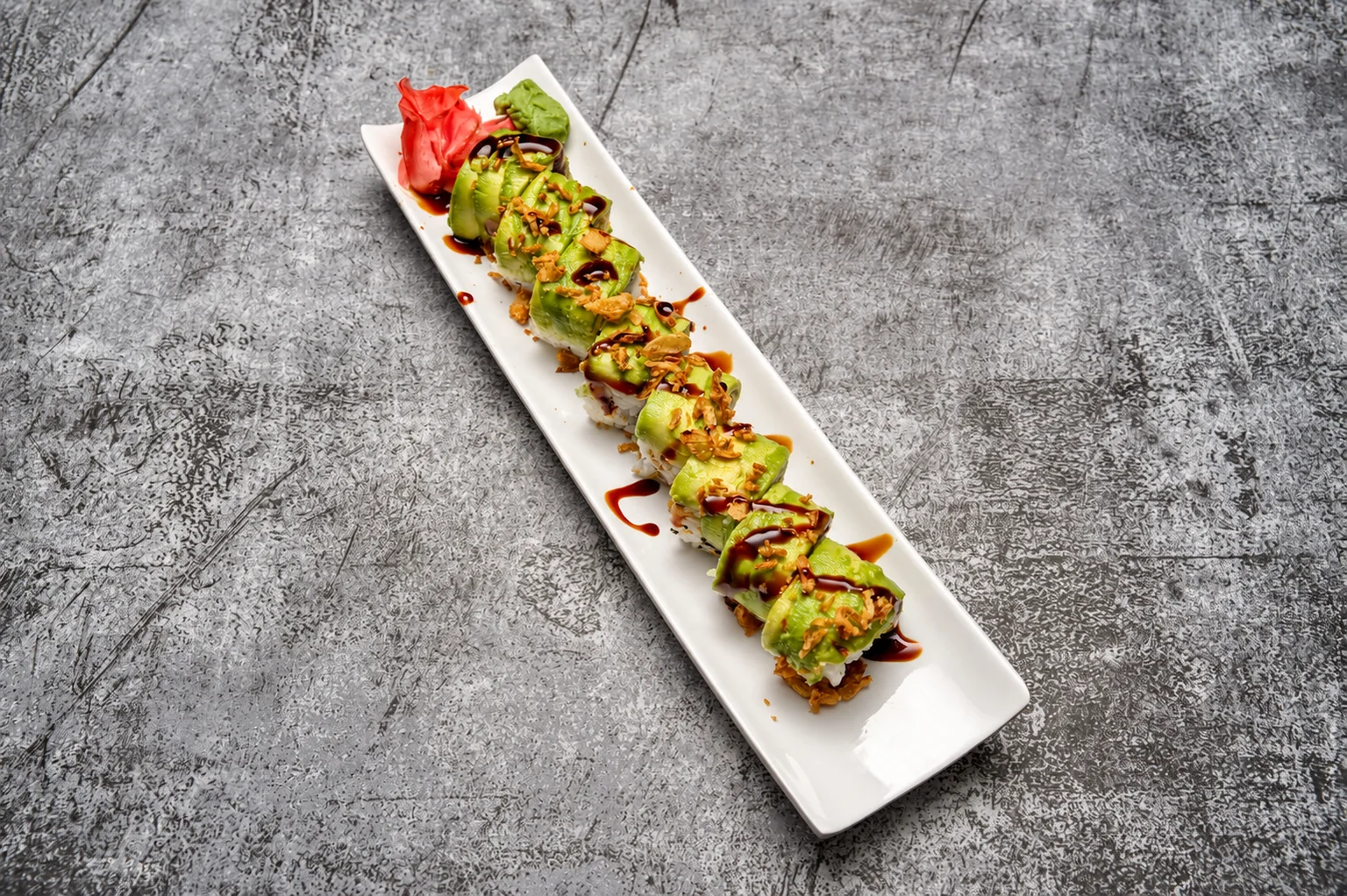 Rolls topped with avocado slices and sauce on a long white plate, grey concrete background.